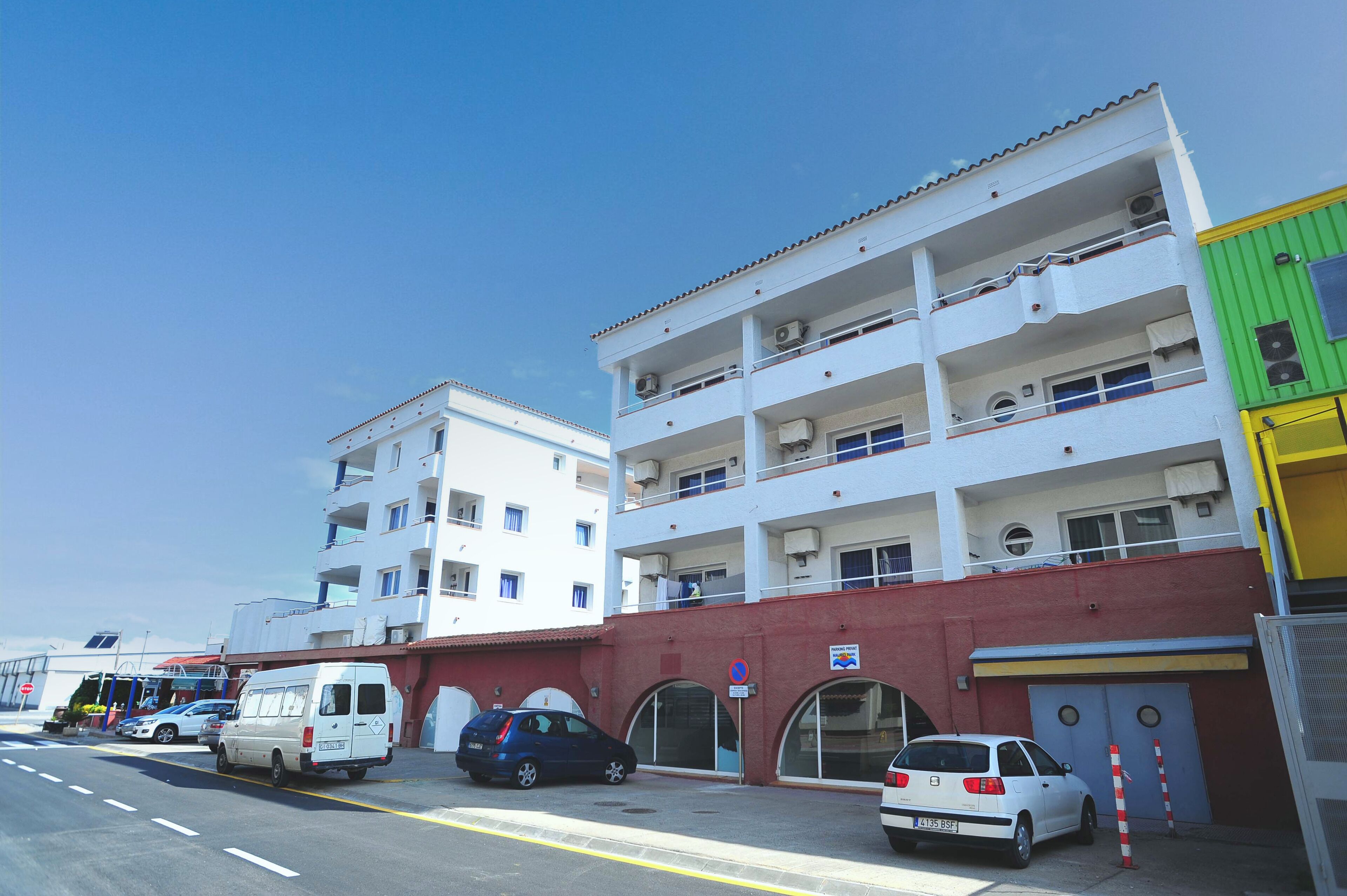 A multi-story building with balconies, a parking area, and a red wall with arched doorways.