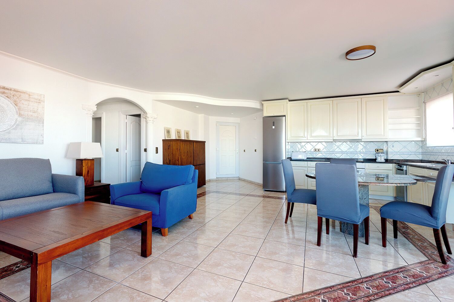 A modern living room with a sofa, armchair, and dining table. A kitchen with white cabinets and a refrigerator is visible in the background.