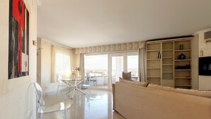 Un vaste espace de vie comprenant une table à manger en verre, des chaises transparentes, un canapé beige et une grande fenêtre donnant sur un paysage agréable.