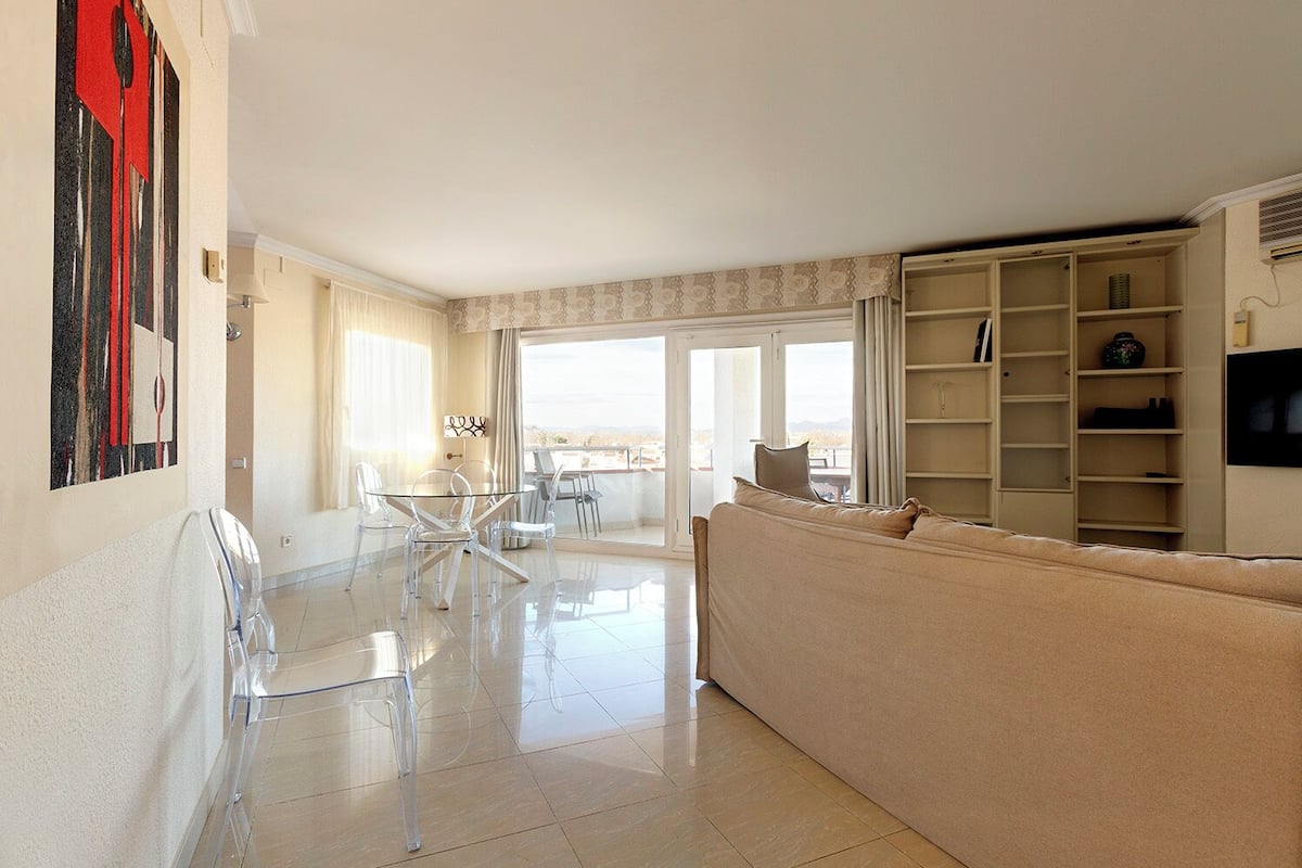 Een ruime woonkamer met een glazen eettafel, transparante stoelen, een beige bank en een groot raam met uitzicht.