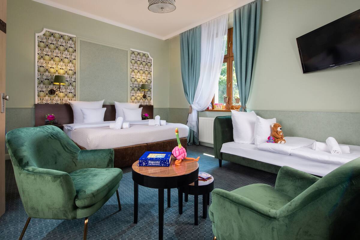 Une chambre d’hôtel avec deux lits, un canapé, des fauteuils verts, une petite table ronde et un téléviseur fixé au mur.