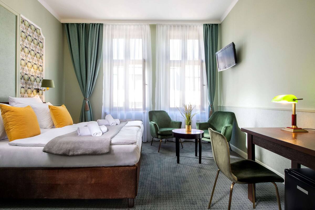 Une chambre d’hôtel comprenant un lit, deux chaises, un bureau, une télévision et une fenêtre avec des rideaux.