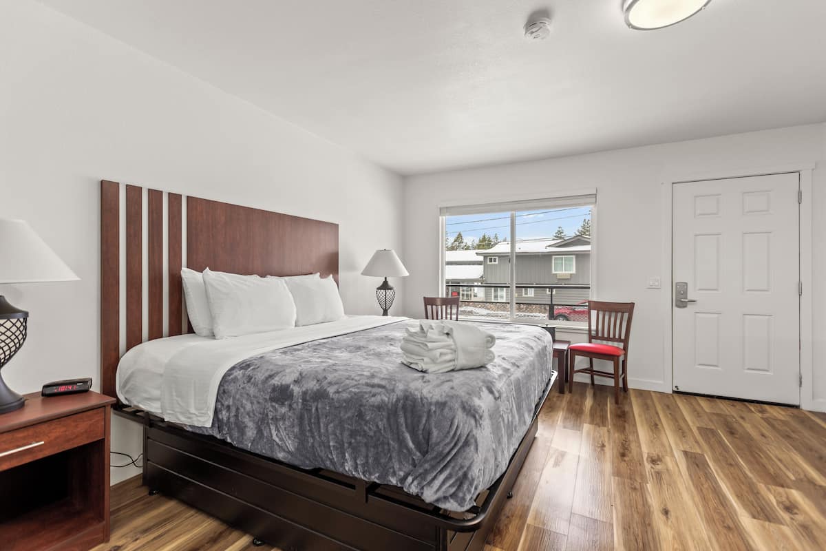 Een hotelkamer met een bed, nachtkastje, lamp, stoel en een raam met uitzicht op andere gebouwen.