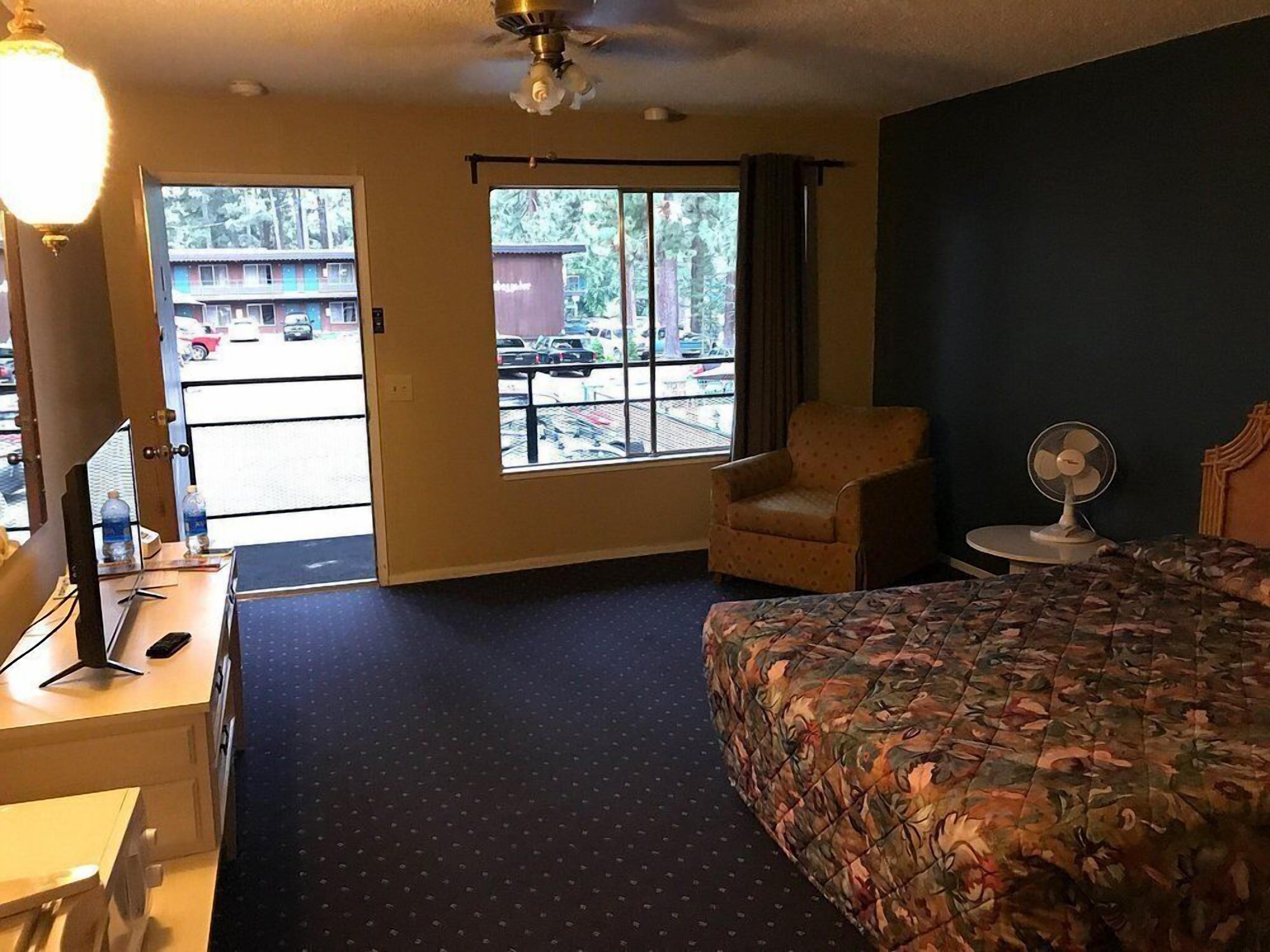 Une chambre d’hôtel comprenant un lit, un bureau avec un ordinateur, une chaise, une fenêtre donnant sur le parking et un ventilateur de plafond.