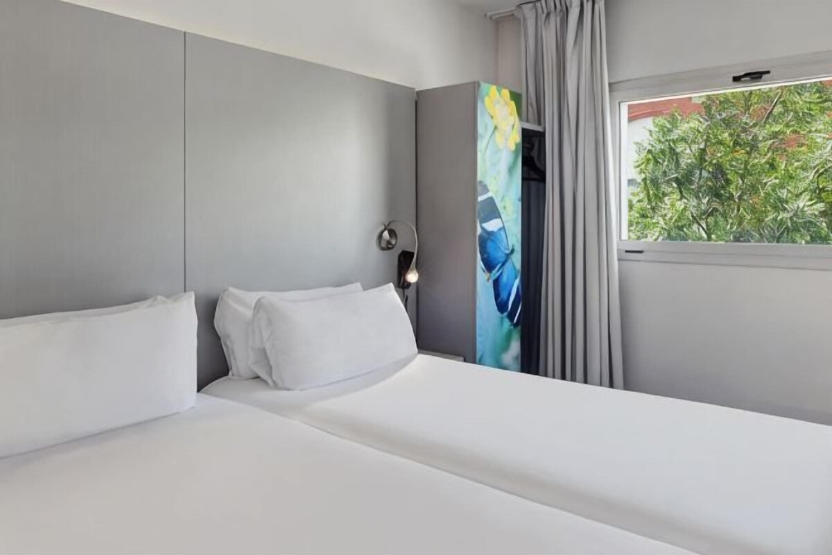 A hotel room with a bed, a nightstand, a painting, and a window with curtains.
