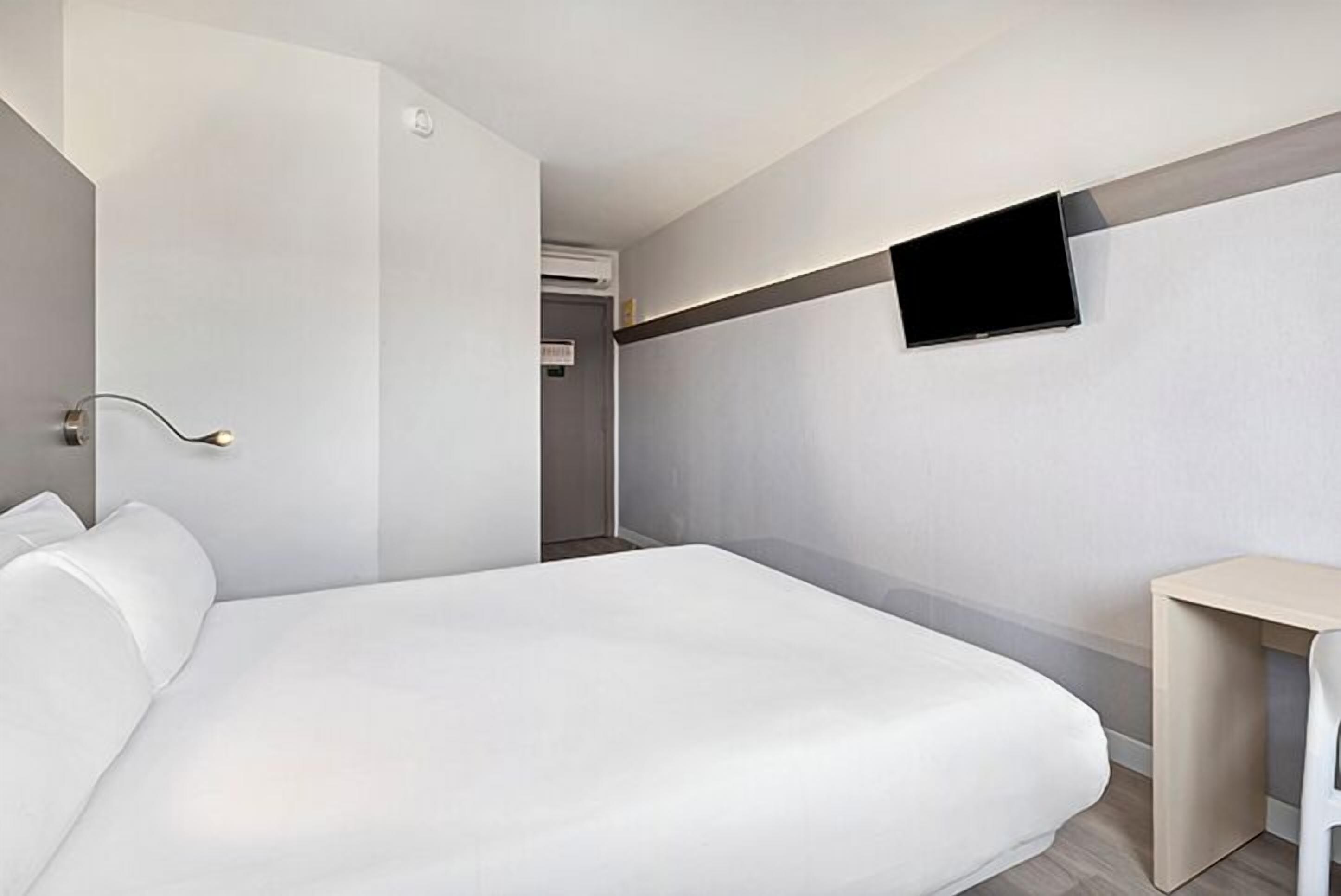 A modern hotel room with a large bed, a flat-screen TV mounted on the wall, and a small bedside table.