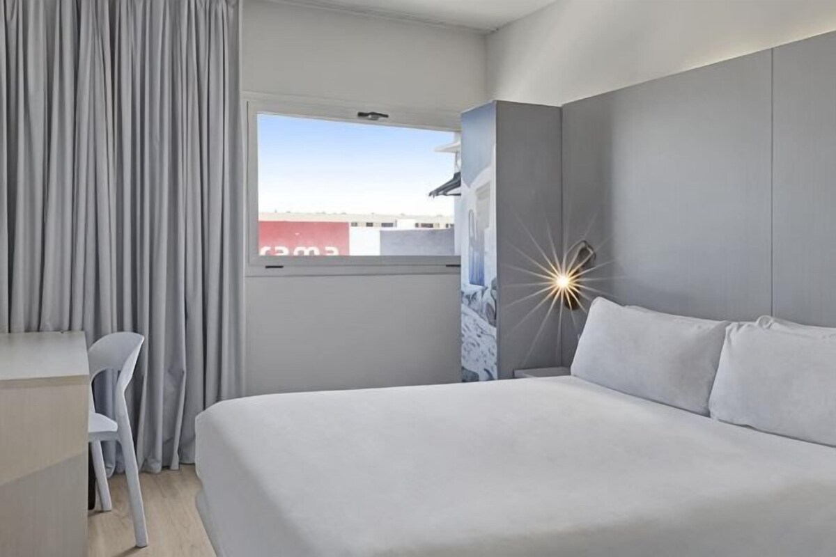 A hotel room with a bed, a desk, a chair, a window with curtains, and a decorative wall piece.