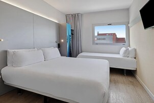 Una habitación de hotel moderna con una cama grande, otra más pequeña, un televisor y un ventanal con vista a la ciudad.
