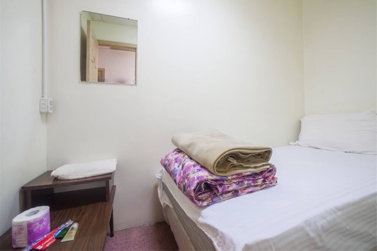 A small, clean room with a bed, a nightstand, a mirror, and a window.