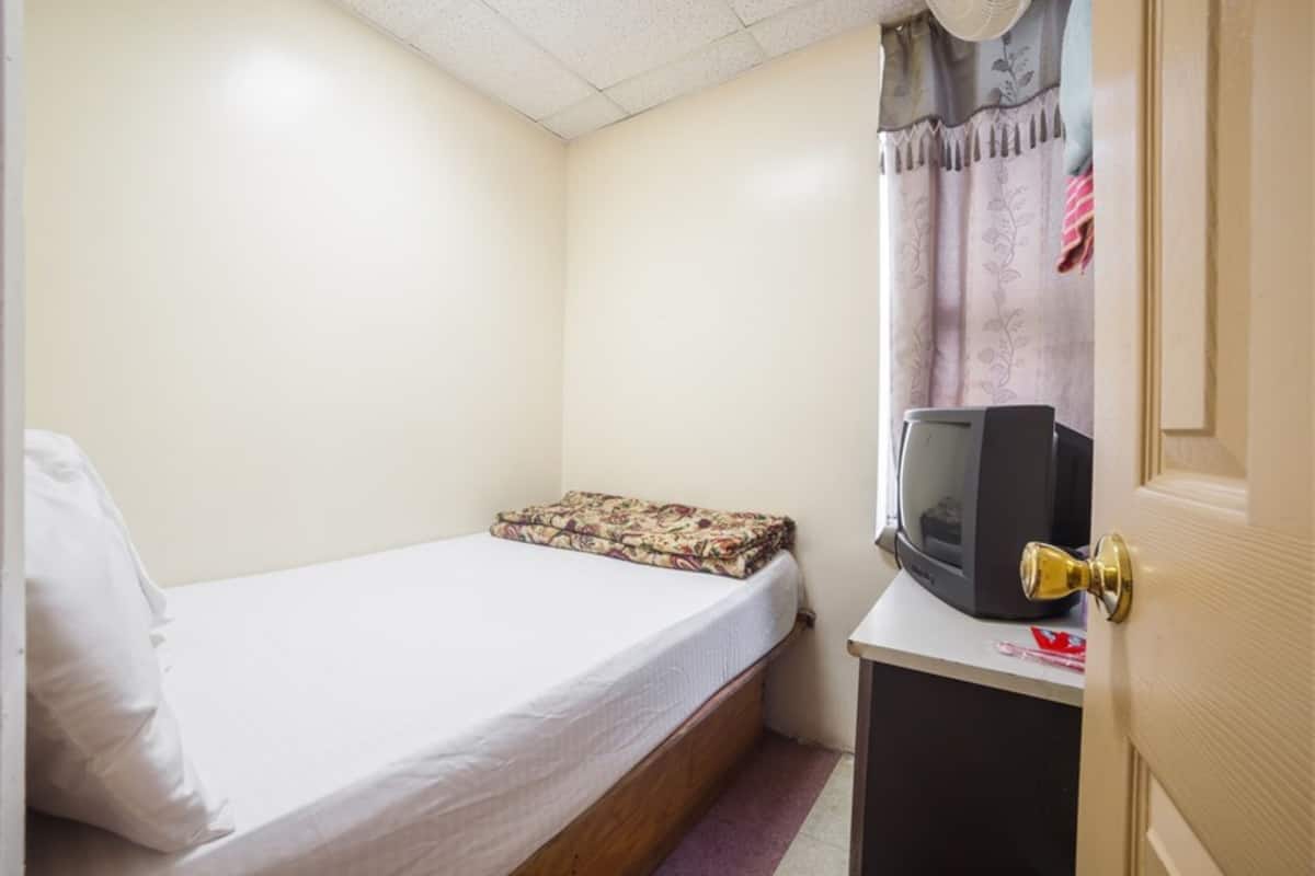 A small, single-bed room with a wooden bed frame, a white mattress, a patterned blanket, a television on a small table, and a door with a golden handle.
