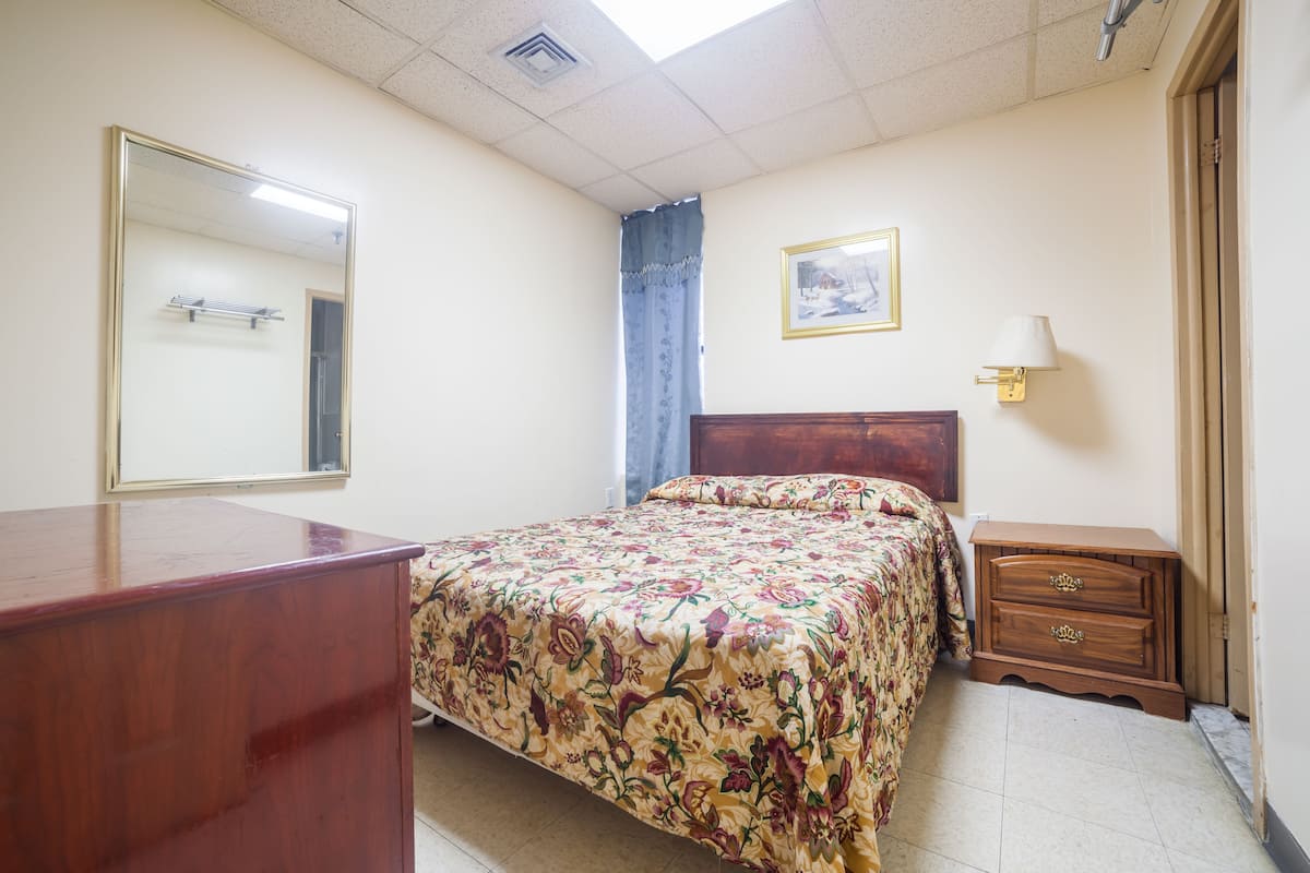 A bedroom with a bed, a wooden dresser, a nightstand, a mirror, a framed picture on the wall, and a window with a curtain.