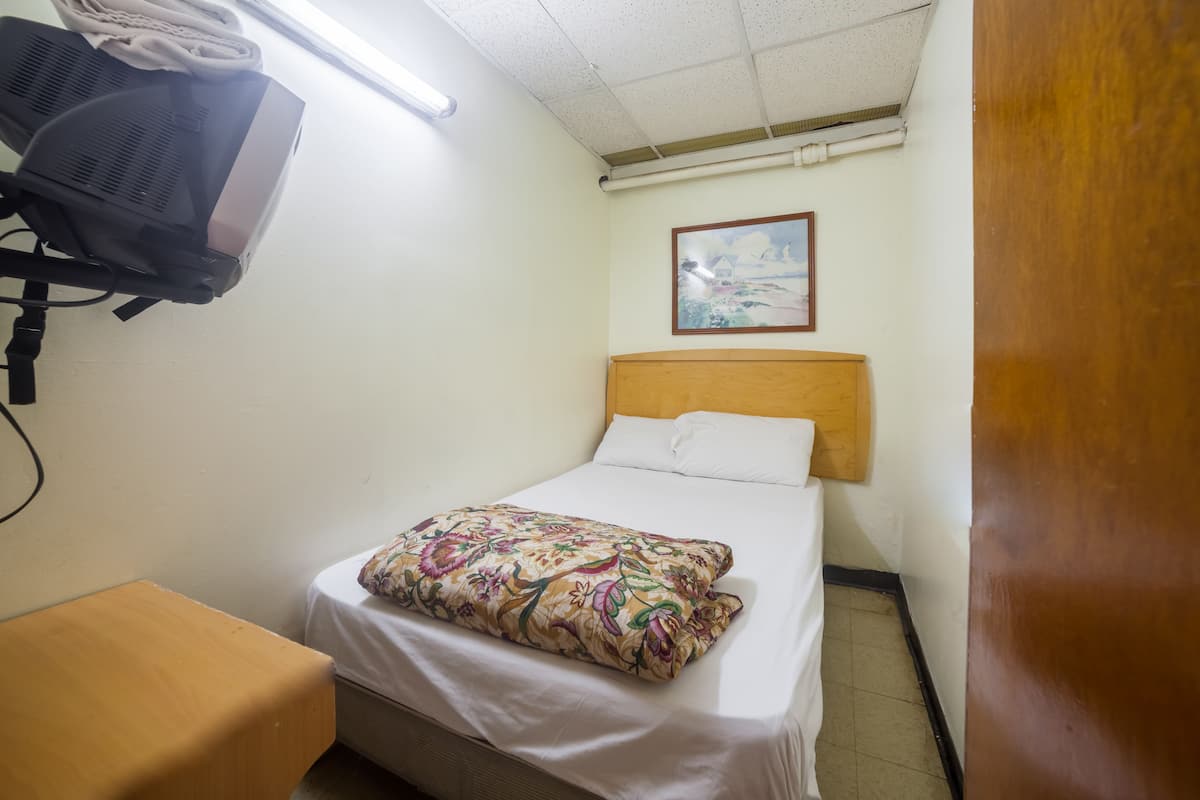 A hotel room with a bed, a wooden headboard, a white pillow, a floral blanket, a small wooden table, and a ceiling fan.