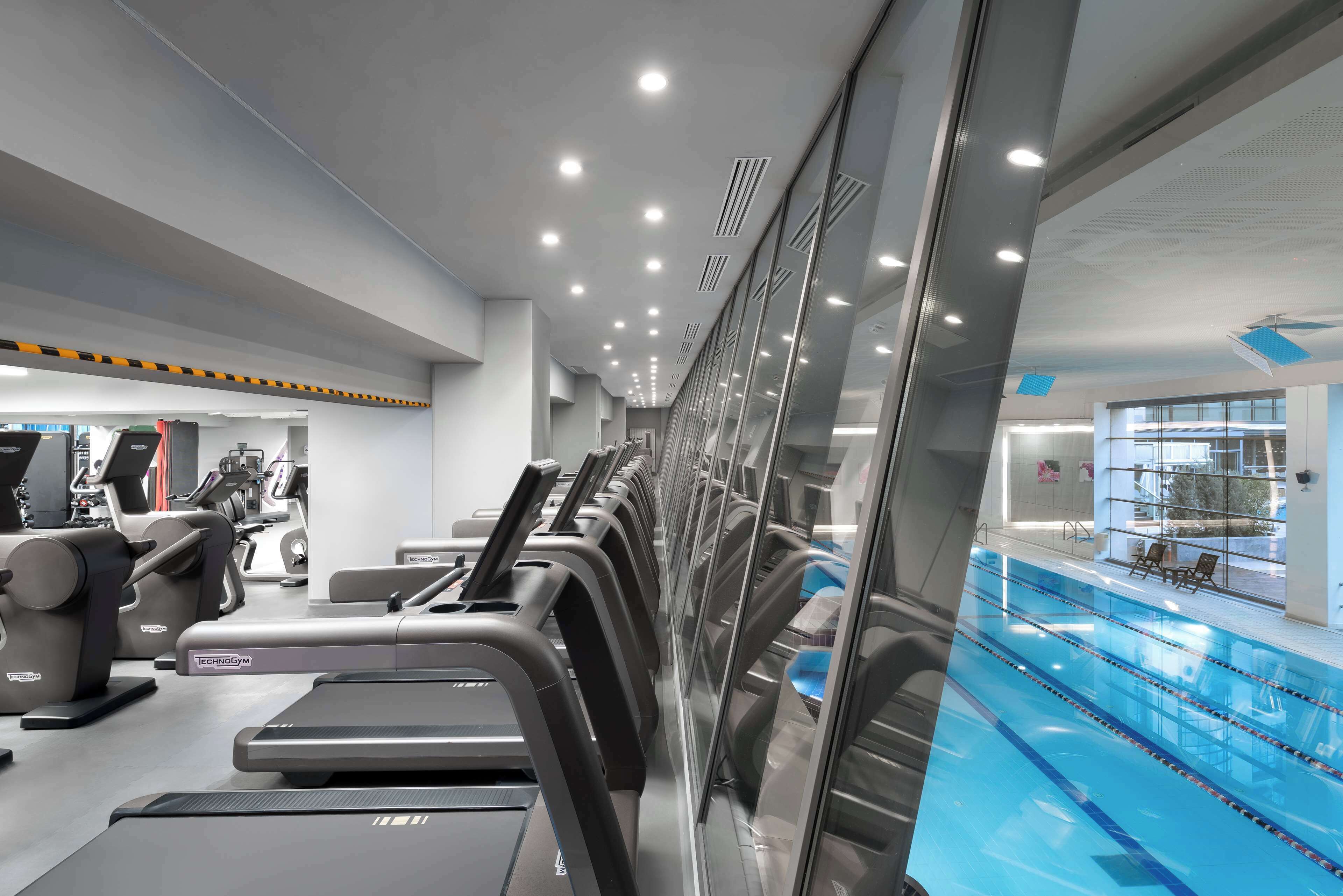 A modern gym with treadmills and exercise equipment, a glass wall reflecting the interior, and a pool area visible through the glass.