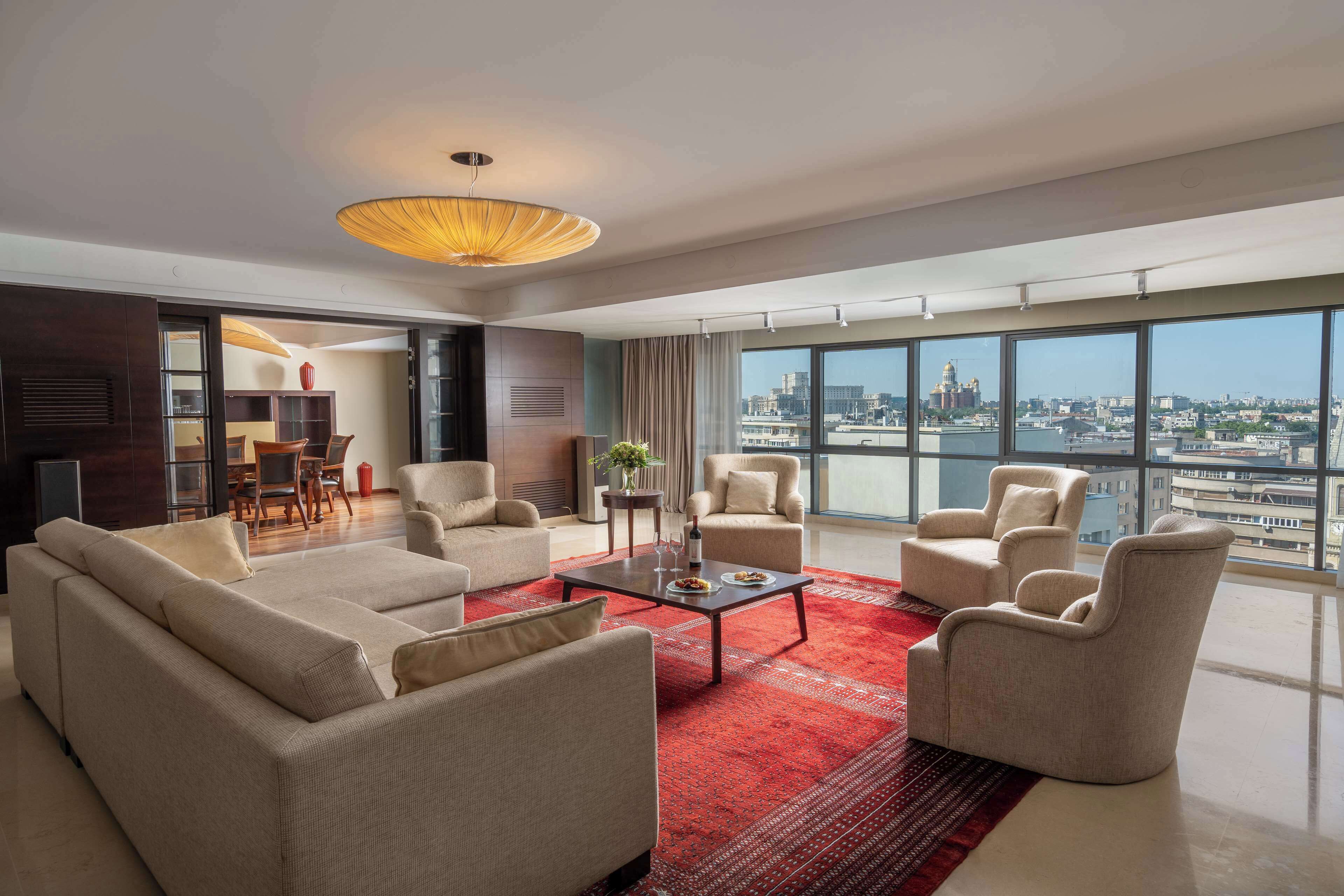 A modern living room with a red rug, beige sofas, and armchairs, a dining area with a table and chairs, and large windows offering a city view.