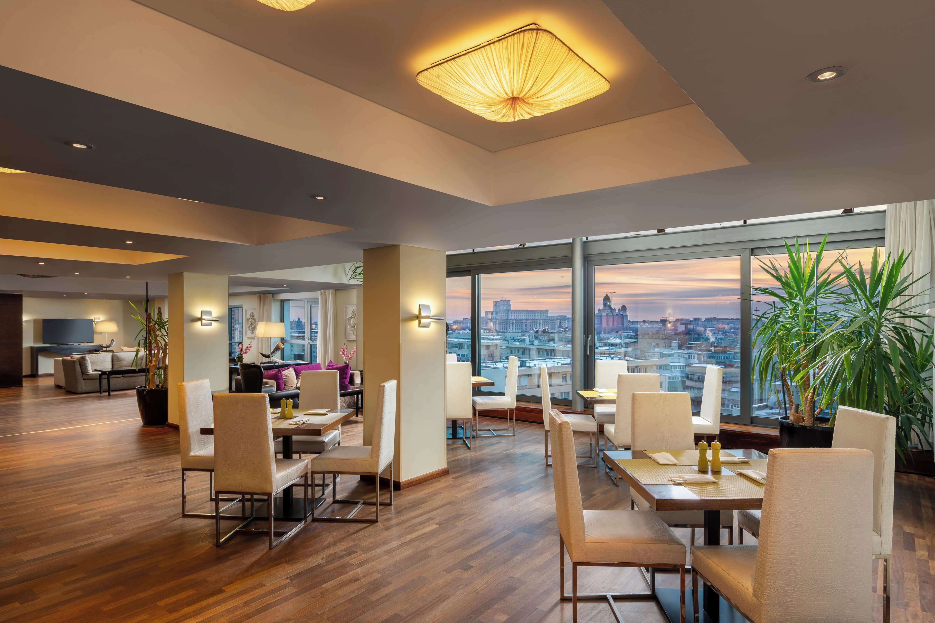 A modern hotel dining area with wooden flooring, white chairs, and tables. Large windows offer a city view.