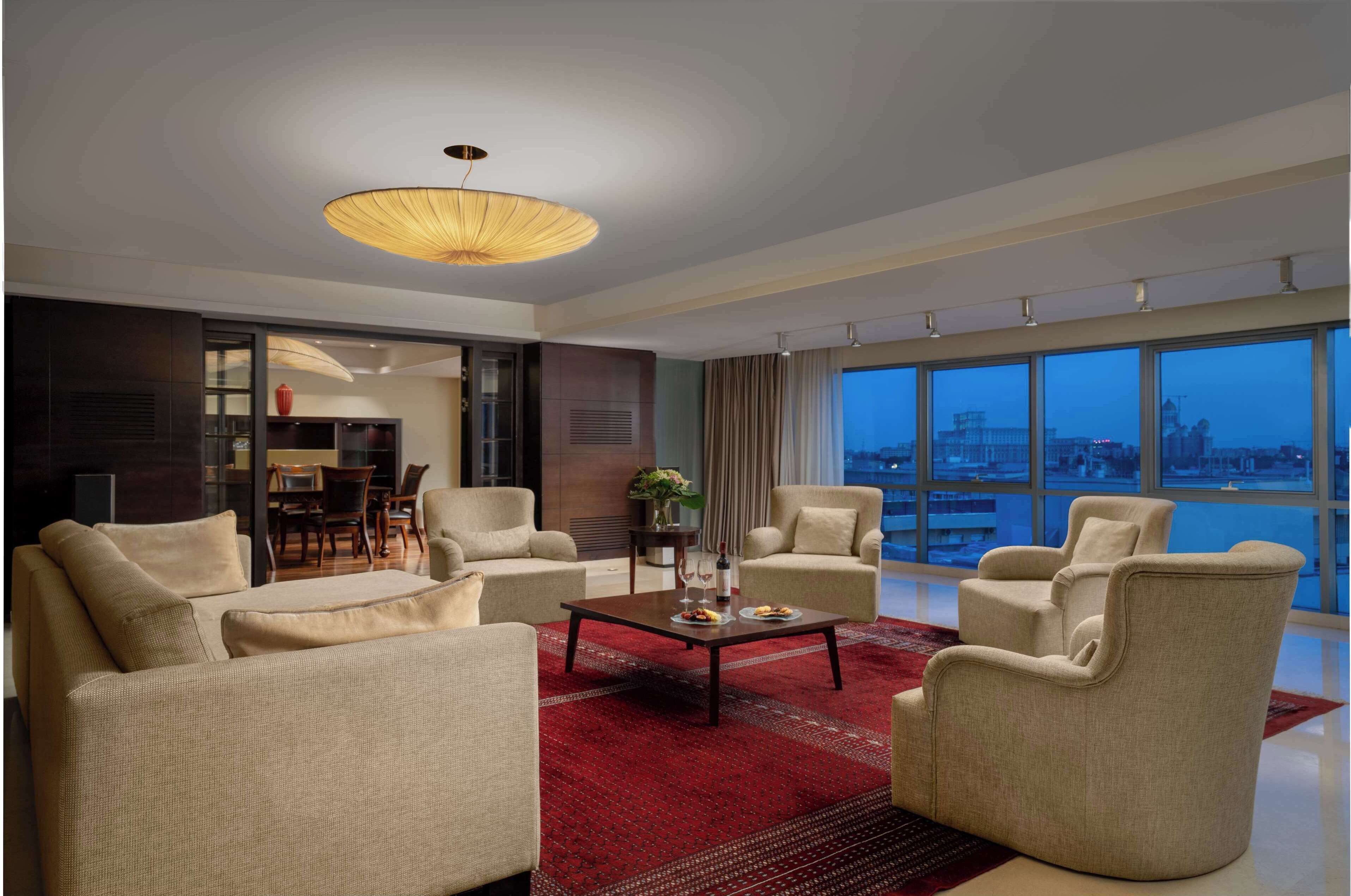 A modern living room with a large window, a red rug, and a dining area visible through an open door.