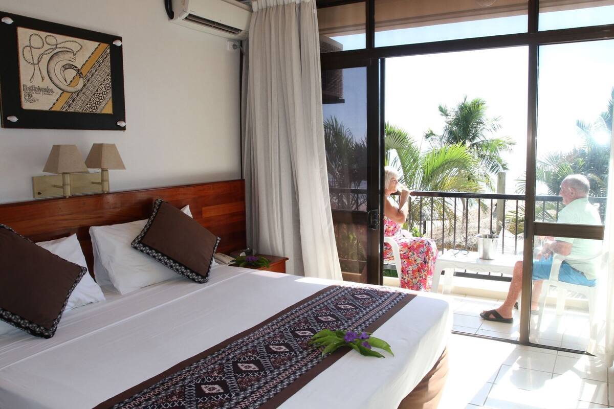 Habitación de hotel con una cama grande, cabecera de madera, lámpara de noche, una obra de arte enmarcada y un balcón con vista a palmeras.