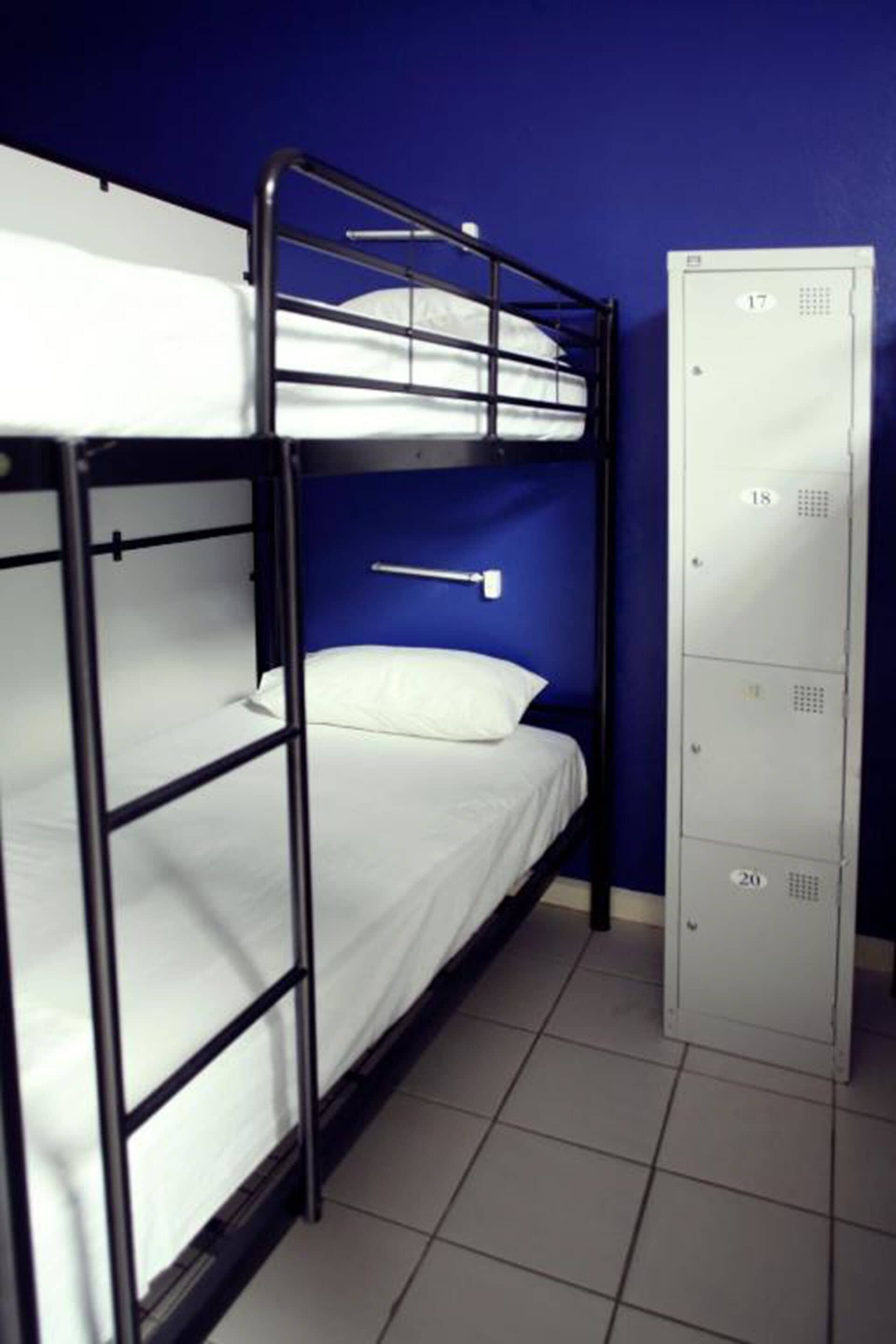 A bunk bed with a white mattress and a metal frame, a single bed with a white mattress and a pillow, and a tall metal storage unit with multiple compartments.