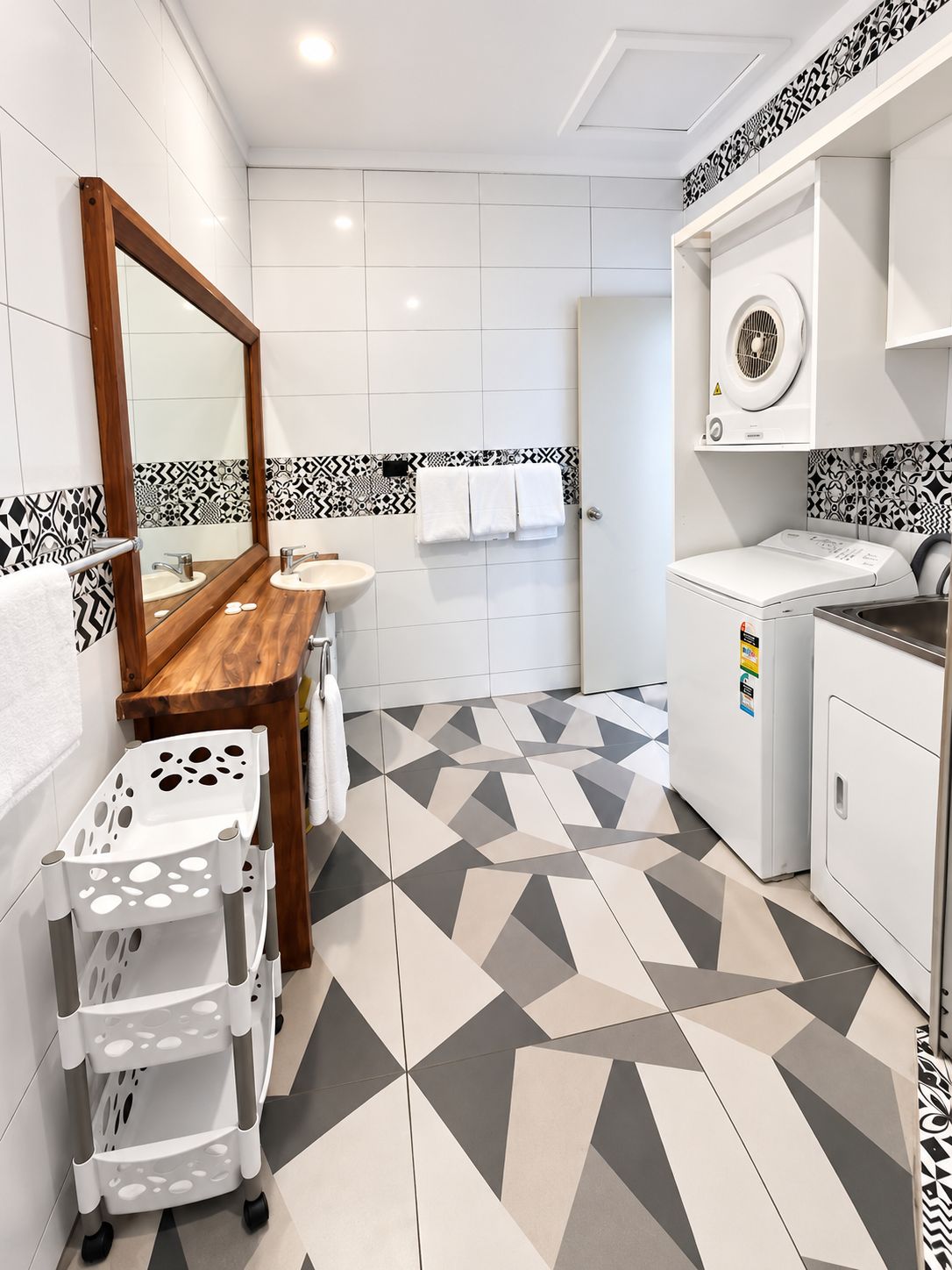 A modern bathroom with a geometric patterned floor, a wooden vanity, a large mirror, a sink, a towel rack, and a washing machine.