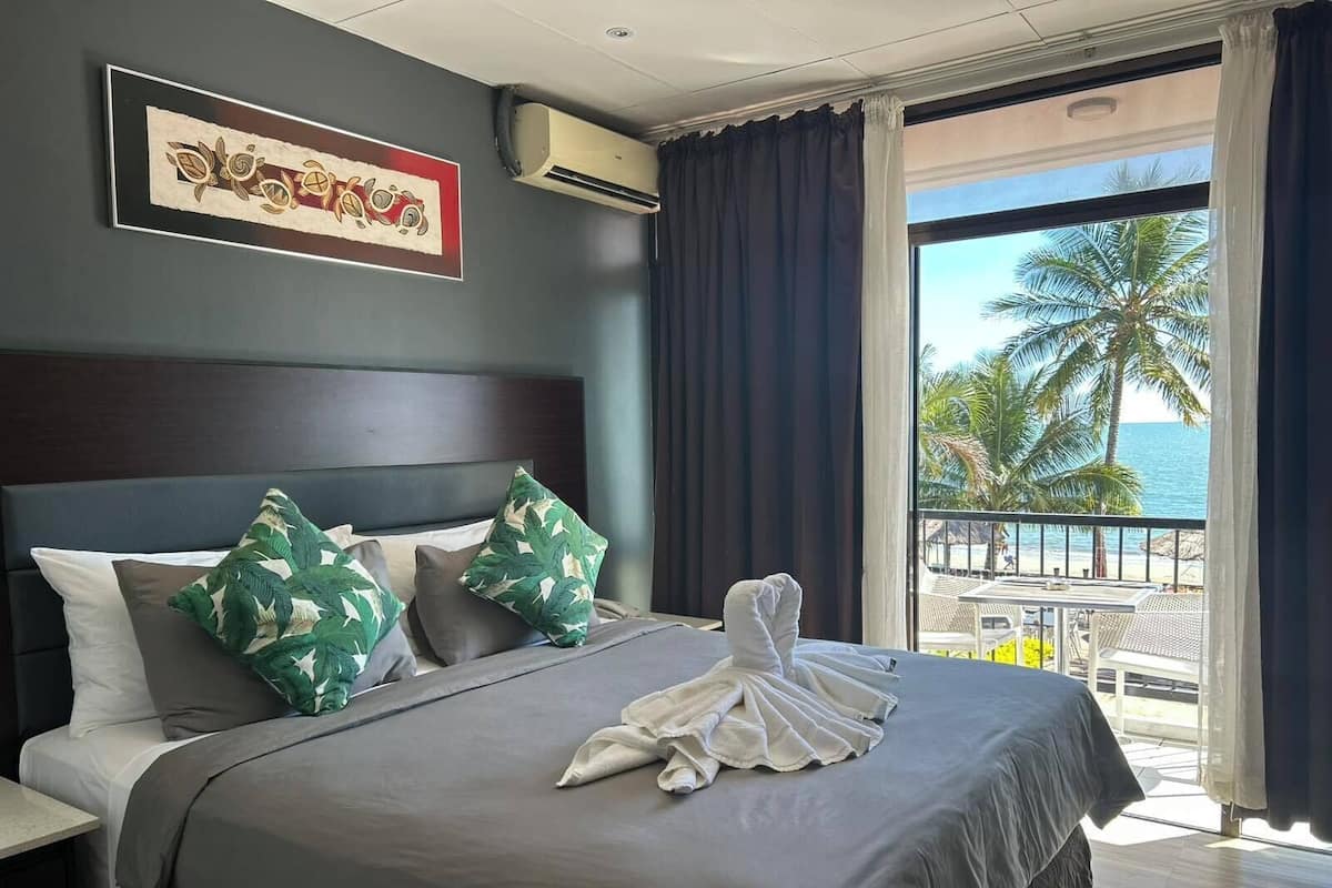 Habitación de hotel con cama, cabecera, una obra de arte enmarcada, unidad de aire acondicionado y balcón con vistas a palmeras y al mar.