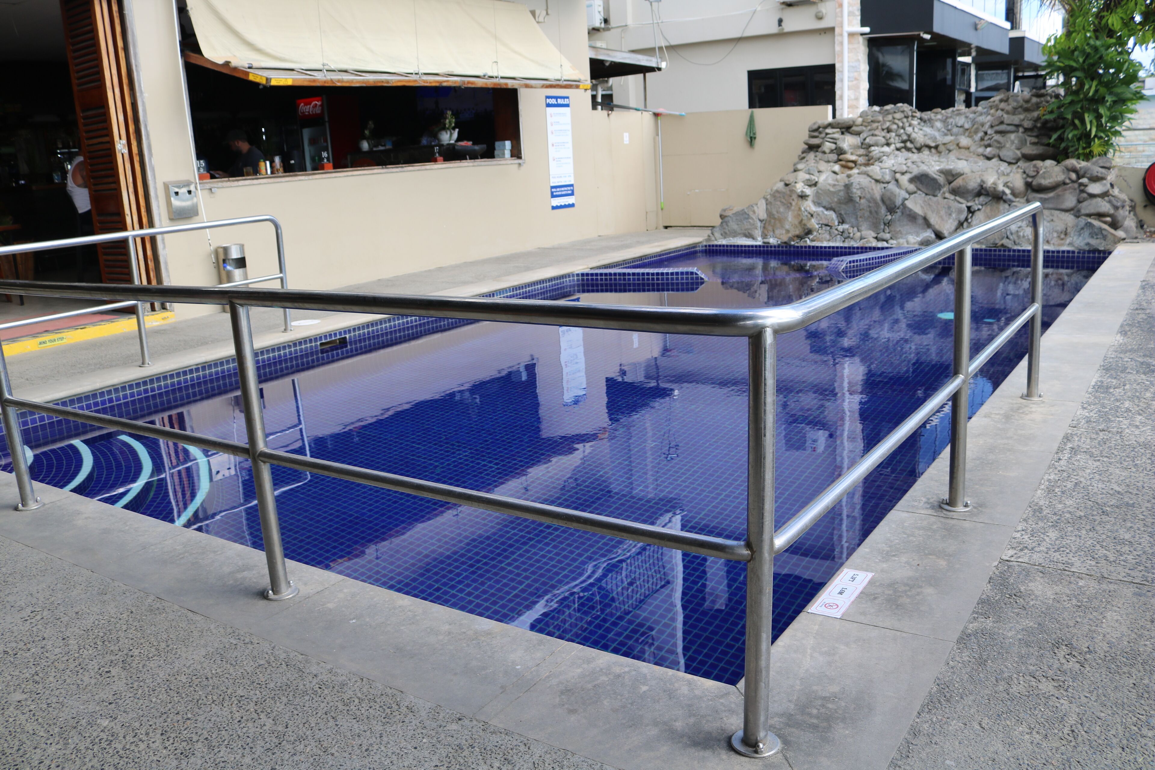 A swimming pool with a metal railing, a rock formation, and a Coca-Cola sign in the background.