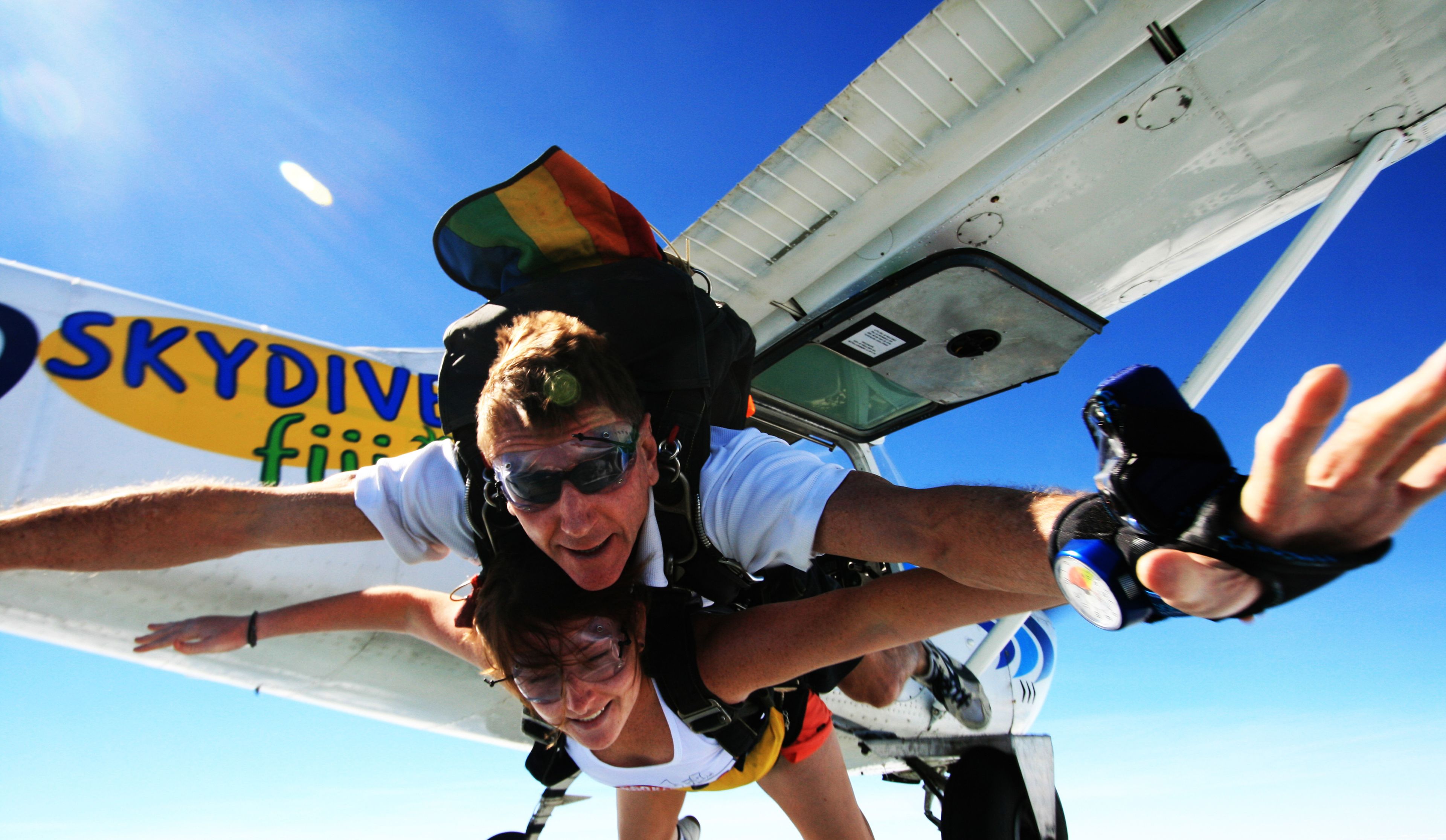 A person skydiving with a parachute, wearing a helmet and goggles, with a clear blue sky in the background.