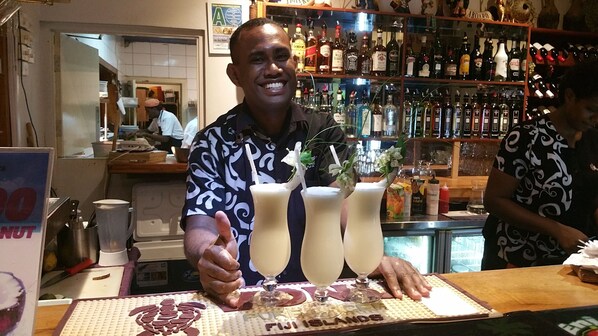 Een bar met verschillende drankflessen op planken, een toonbank met een menukaart en een persoon die een duimpje omhoog steekt.