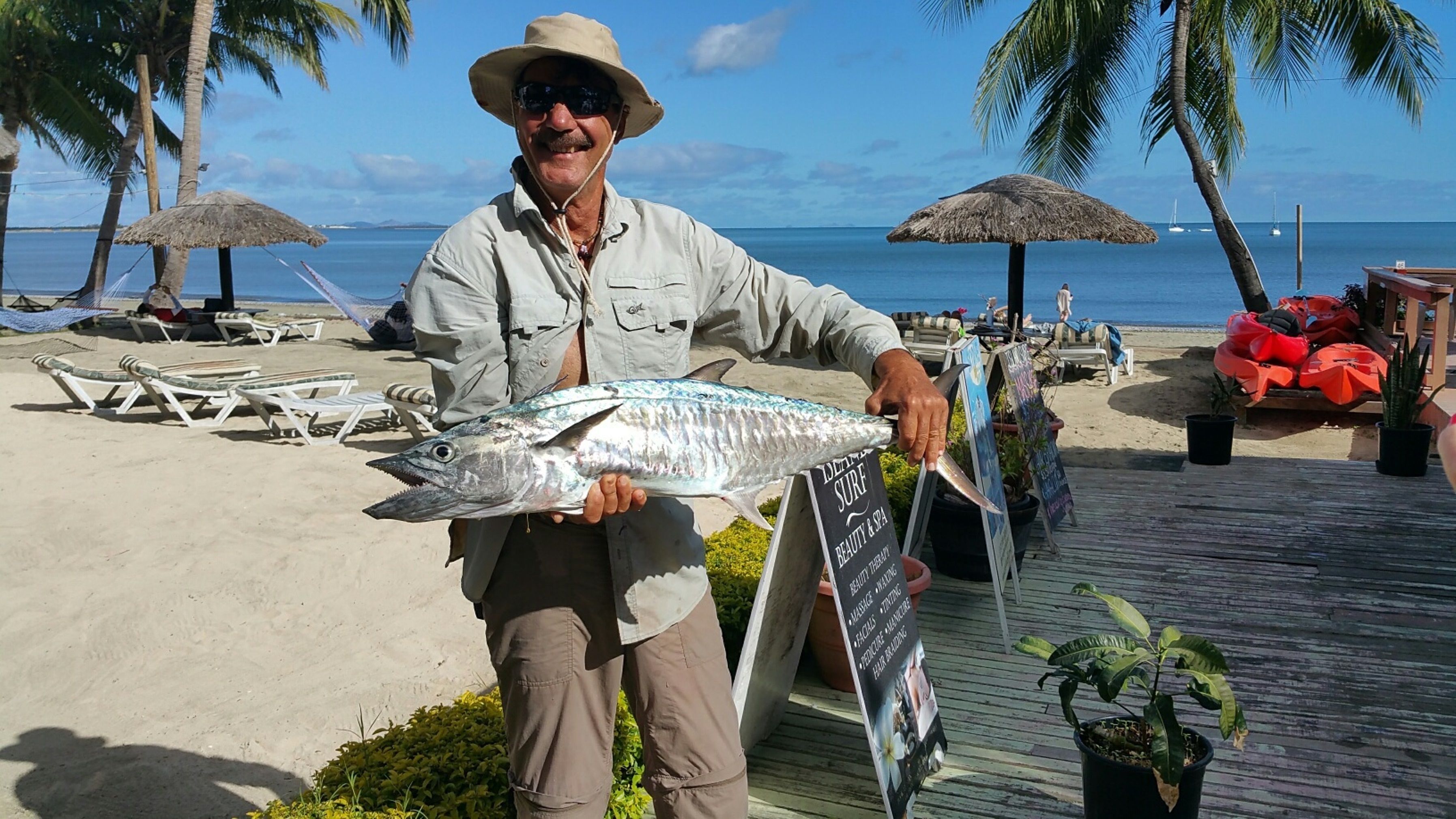 A man holding a fish on a beach with palm trees and lounge chairs.