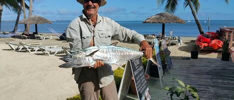 Un homme tenant un poisson sur une plage, avec des palmiers et des chaises longues.