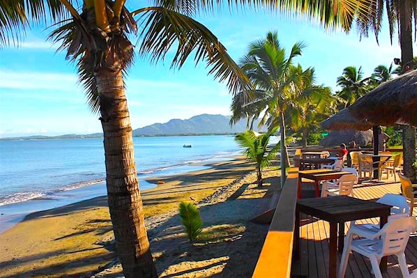 Een restaurant aan het strand met terras, palmbomen en uitzicht op de oceaan.