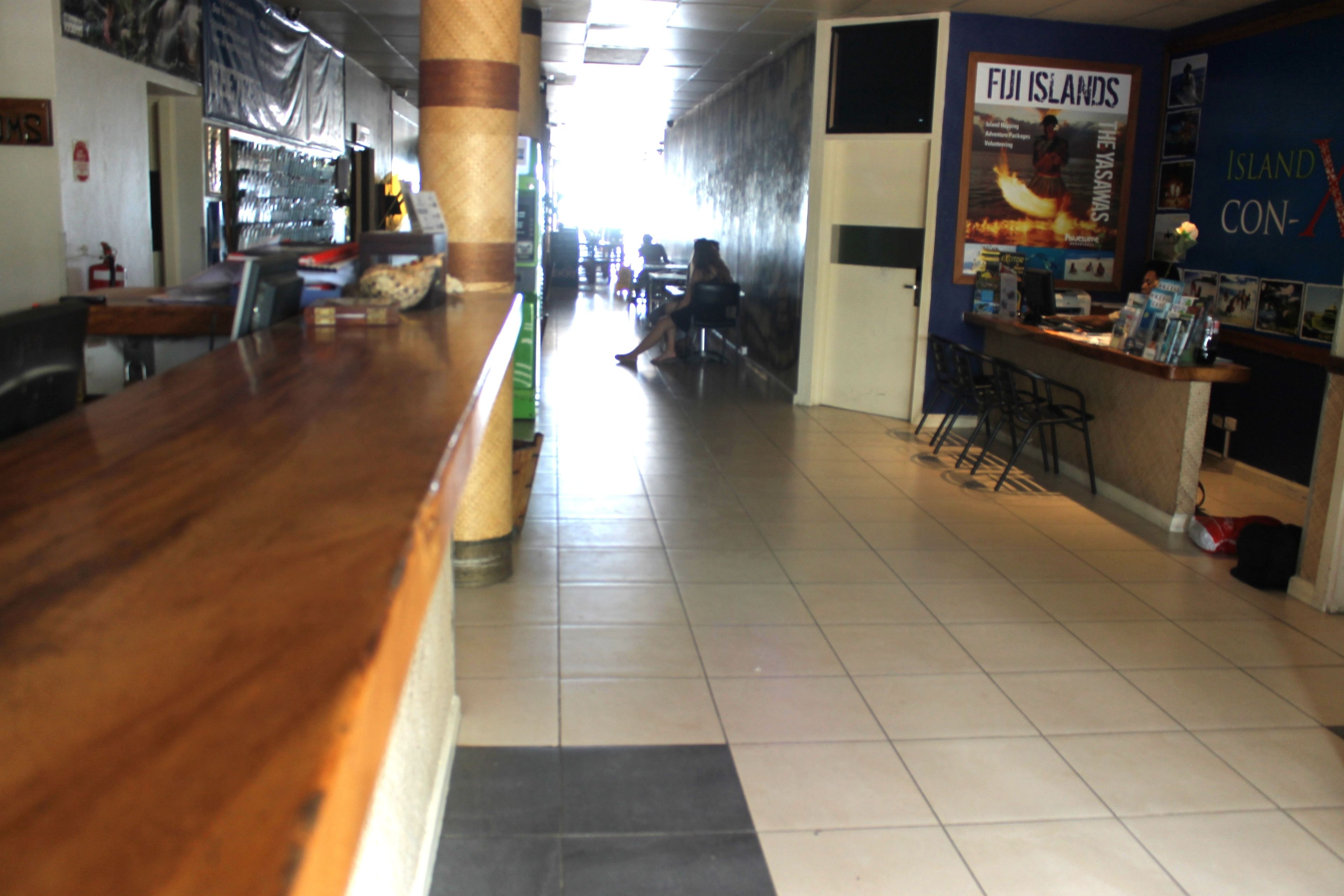 A hotel lobby with a wooden reception desk, a poster advertising Fiji Islands, and a tiled floor.