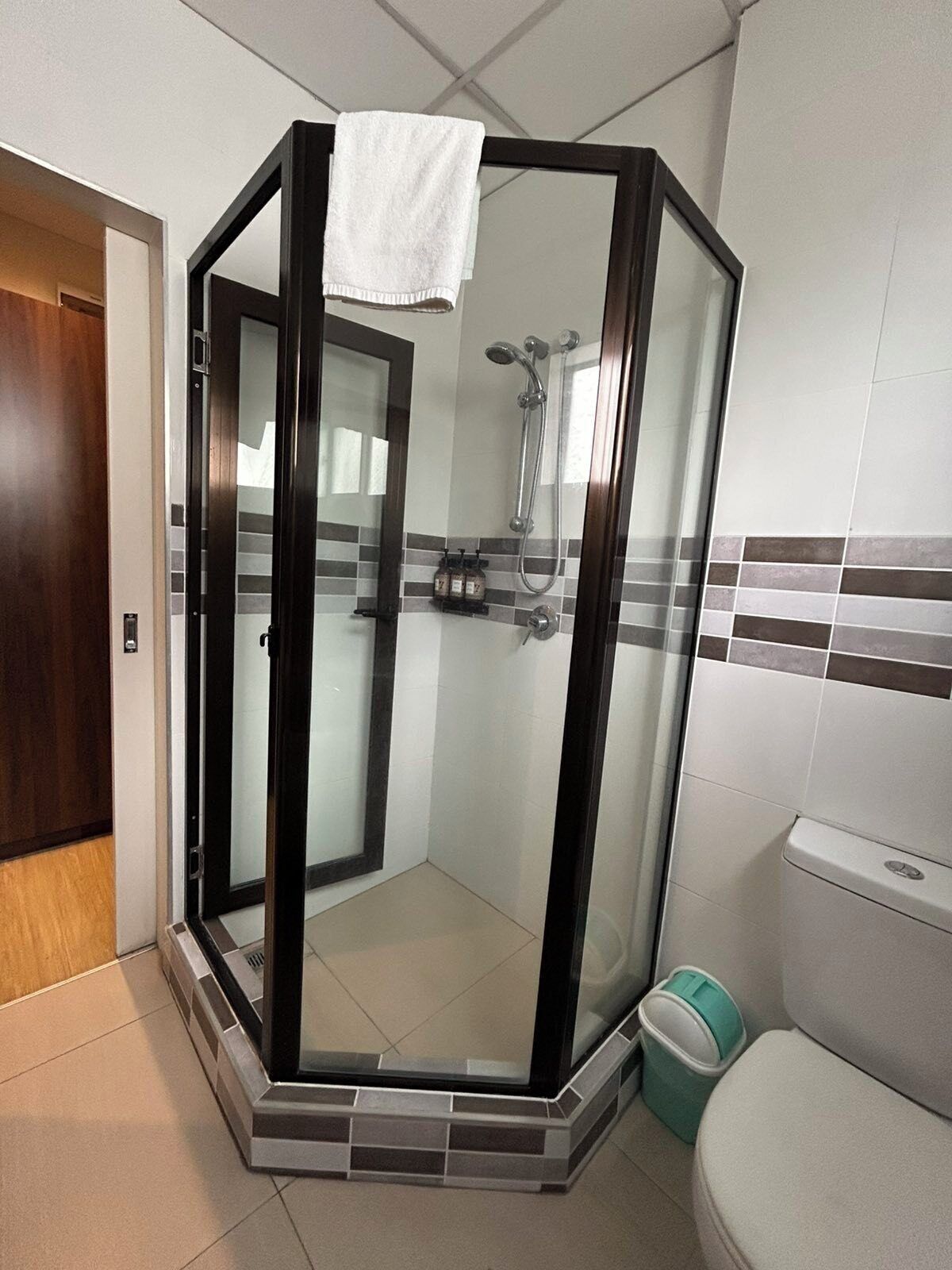 A glass-enclosed shower stall with a white towel hanging on the door, a toilet, and a trash bin in a bathroom with tiled walls and a wooden door.
