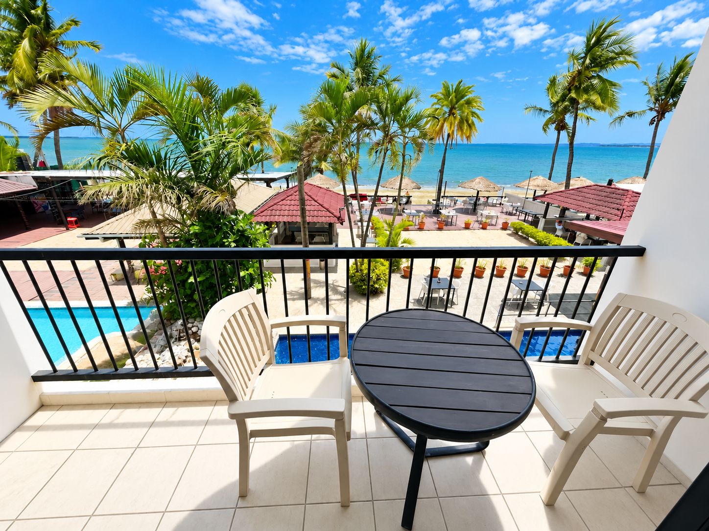 Un balcón con piscina, dos sillas y una mesa con vista a una playa con palmeras y cielo despejado.