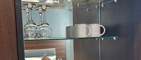 A hotel room with a minibar, a bed, a glass shelf with wine glasses and mugs, and a Sonifer kettle.
