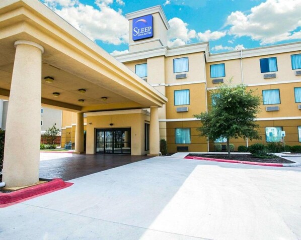 Entrada de un hotel con un letrero que dice "Sleep Inn & Suites", que cuenta con un porche cubierto, alfombra roja y un área ajardinada con un árbol.