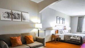 A hotel room with a grey sofa, a wooden coffee table, a bed with white bedding, and a red carpet. - Sleep Inn & Suites New Braunfels (New Braunfels)