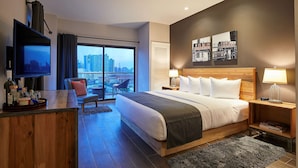A modern hotel room with a large bed, a TV, a desk with a tray of toiletries, a chair, and a view of the city skyline through a large window.