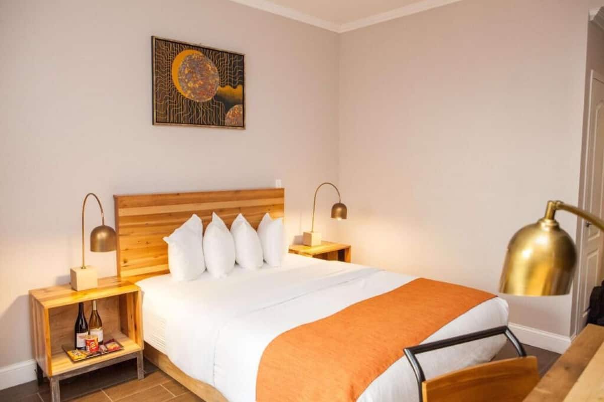 Een hotelkamer met een houten hoofdbord, wit beddengoed, een enkel bed, twee nachtkastjes met lampen, een klein plankje met boeken en flessen, en een schilderij aan de muur.