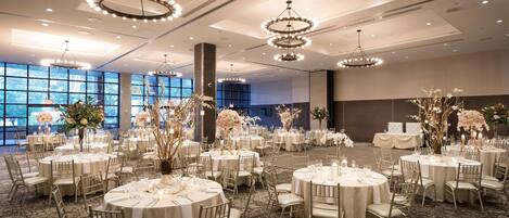 A large, well-lit banquet hall with round tables set for events, featuring elegant chandeliers and floral centerpieces.