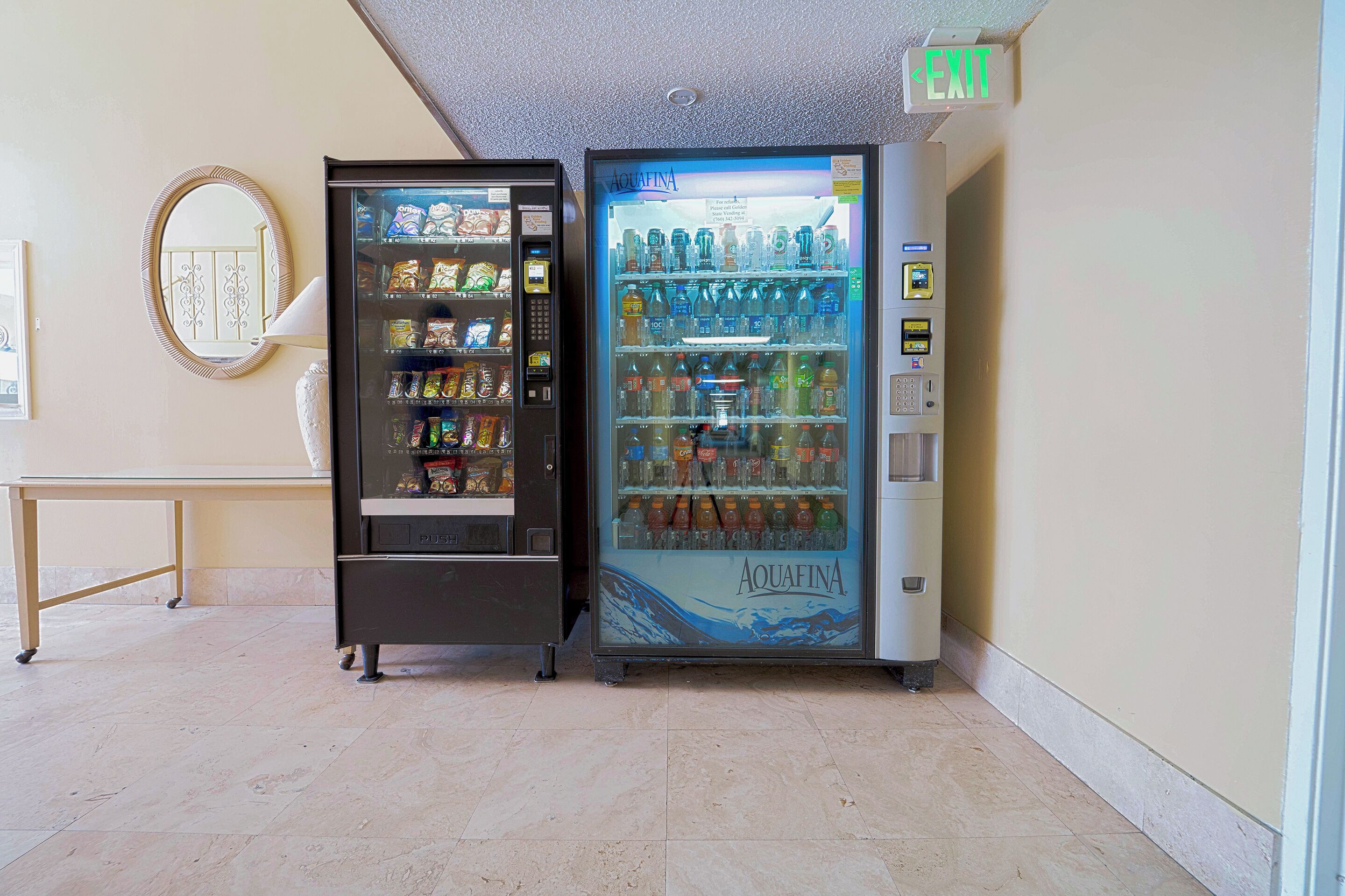 Two vending machines in a hallway with a mirror on the wall and a table.