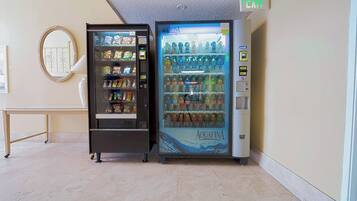 Two vending machines in a hallway with a mirror on the wall and a table.