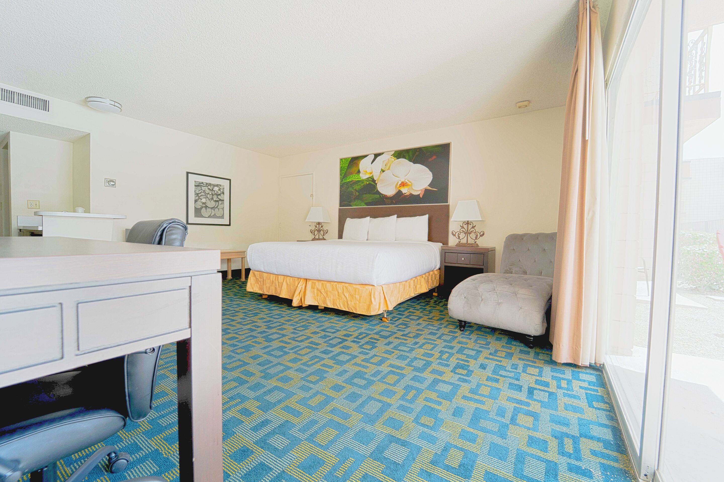 A hotel room with a bed, a desk with a chair, a small table, a lamp, and a window with curtains.