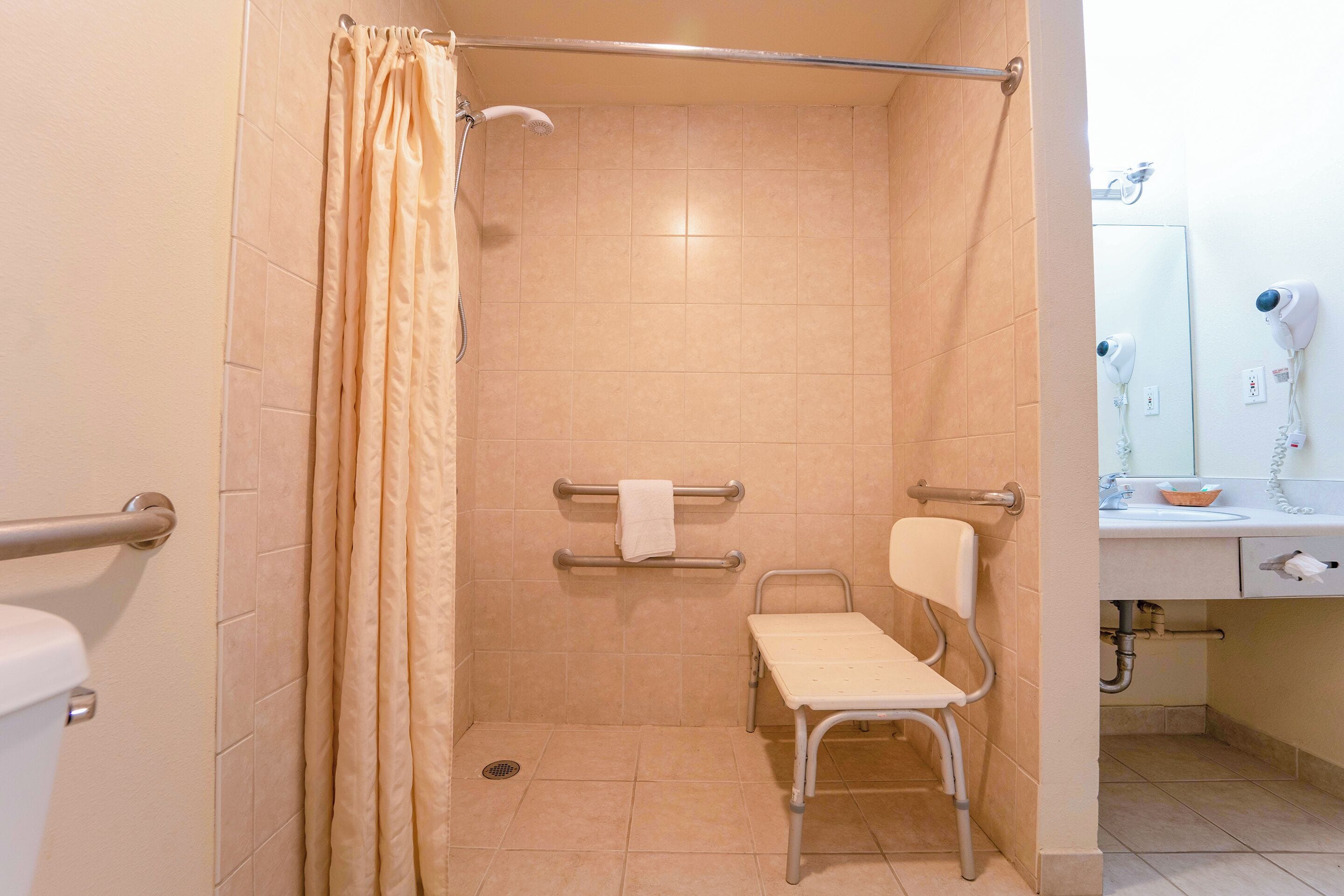 A bathroom with a shower, a toilet, a grab bar, a chair, and a sink with a mirror.