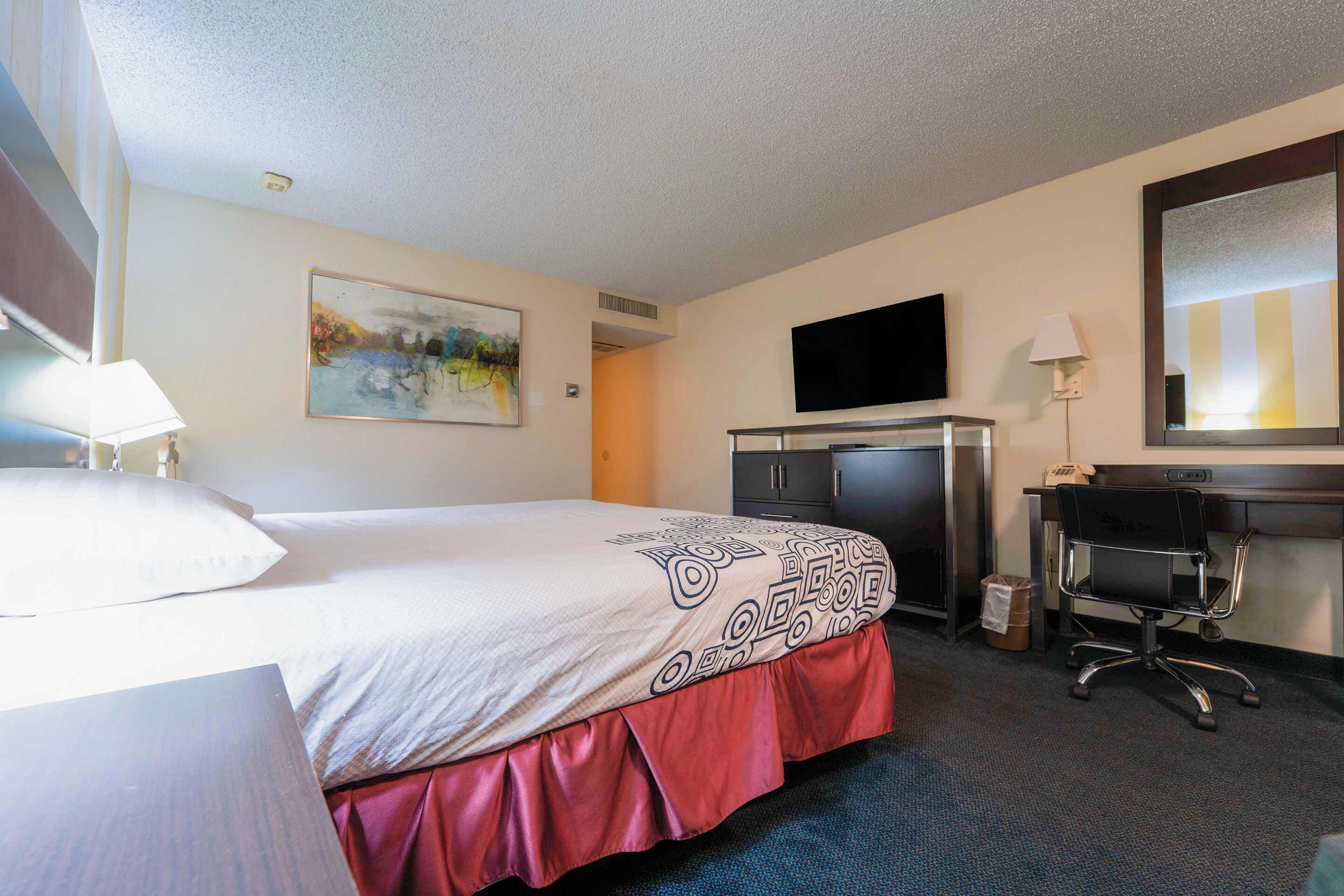 A hotel room with a bed, a desk, a chair, a TV, and a painting on the wall.