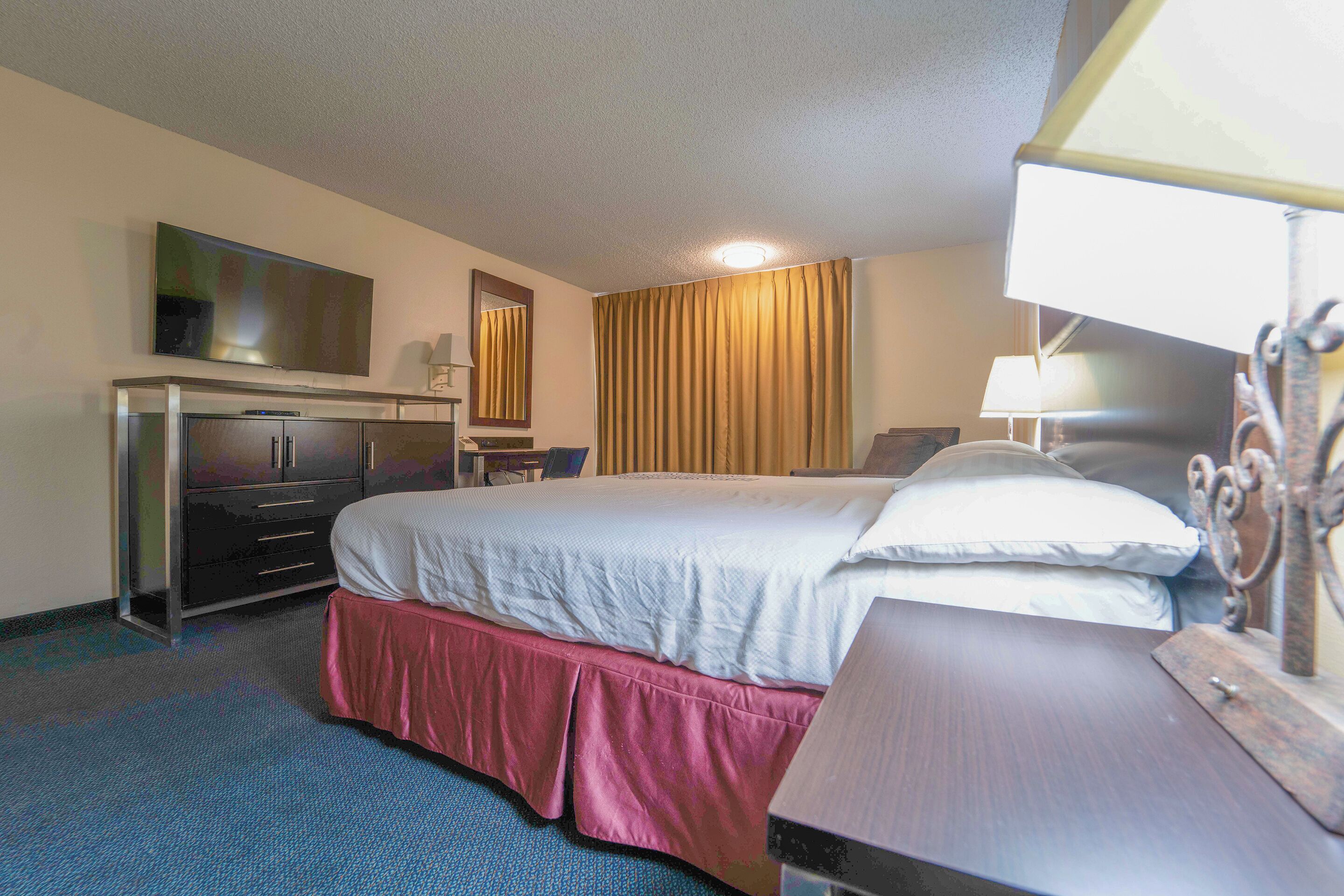 Habitación de hotel con cama, escritorio, televisor, espejo y ventana con cortinas.