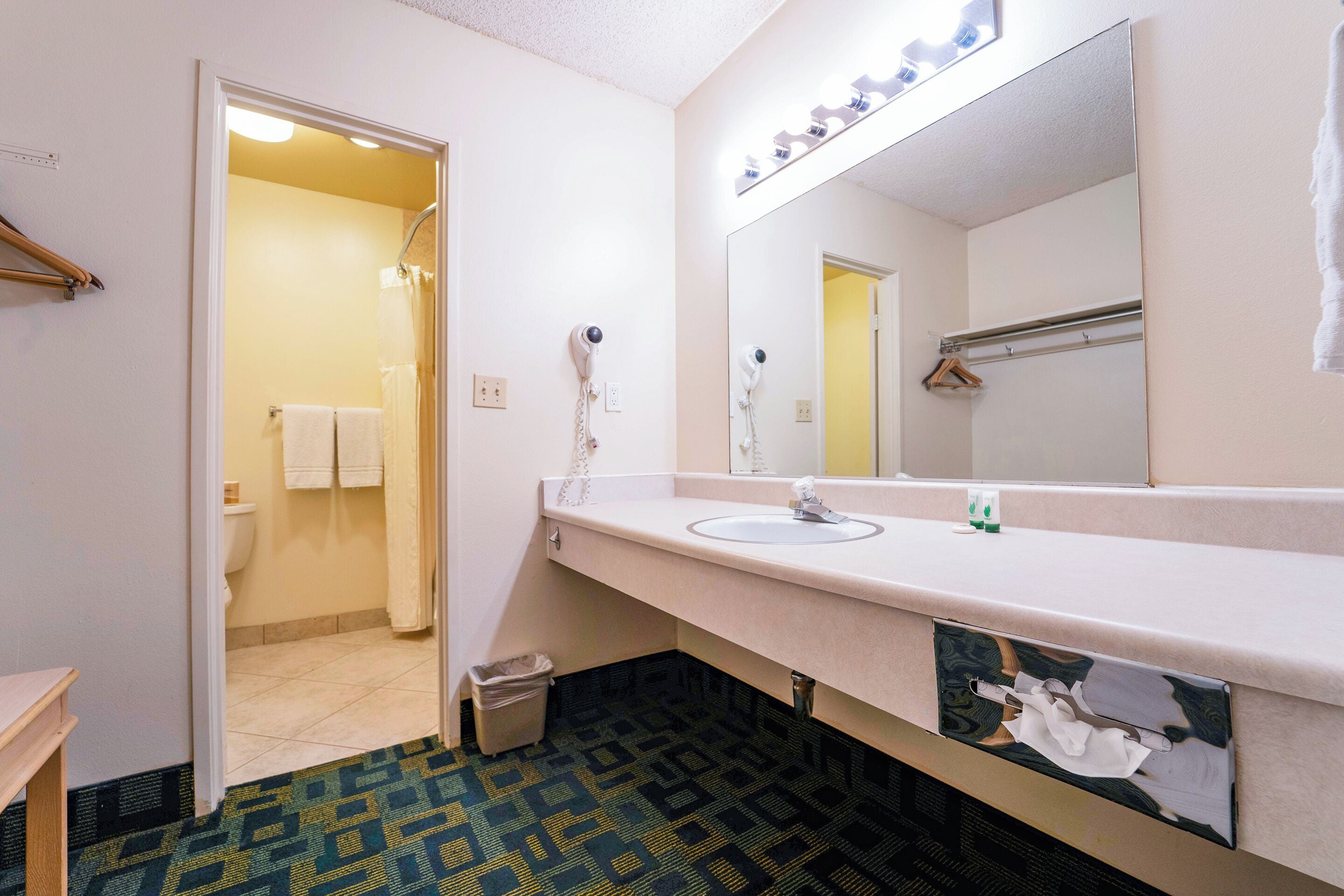 A bathroom with a sink, mirror, and hairdryer. There is a towel rack and a trash can.
