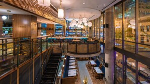 Un bar moderno con una distribución circular de asientos, una escalera que conduce a un nivel superior y vistas de la ciudad a través de amplias ventanas.
