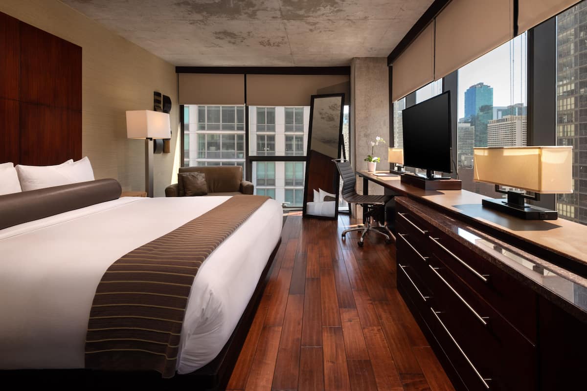 Une chambre d’hôtel dotée d’un grand lit, d’un bureau avec un téléviseur à écran plat, d’une chaise et offrant une vue sur la ville grâce à de grandes fenêtres.