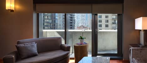 Una sala de estar moderna con un sofá de cuero marrón, una pequeña mesita de madera con una planta y un gran ventanal corredero que da a un balcón con vistas a la ciudad.