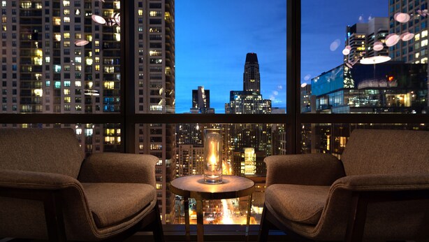 Una habitación con dos sillas y una mesita, un ventanal grande con vistas a la ciudad de noche.